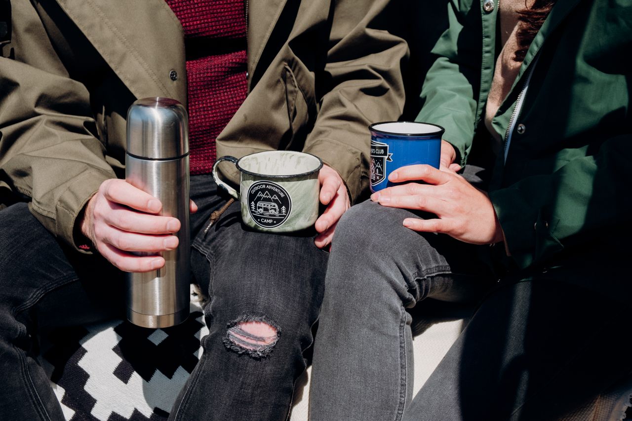 hero-img-01 Casual outdoor moment with a couple holding mugs and a thermos, wearing casual jackets and denim jeans.