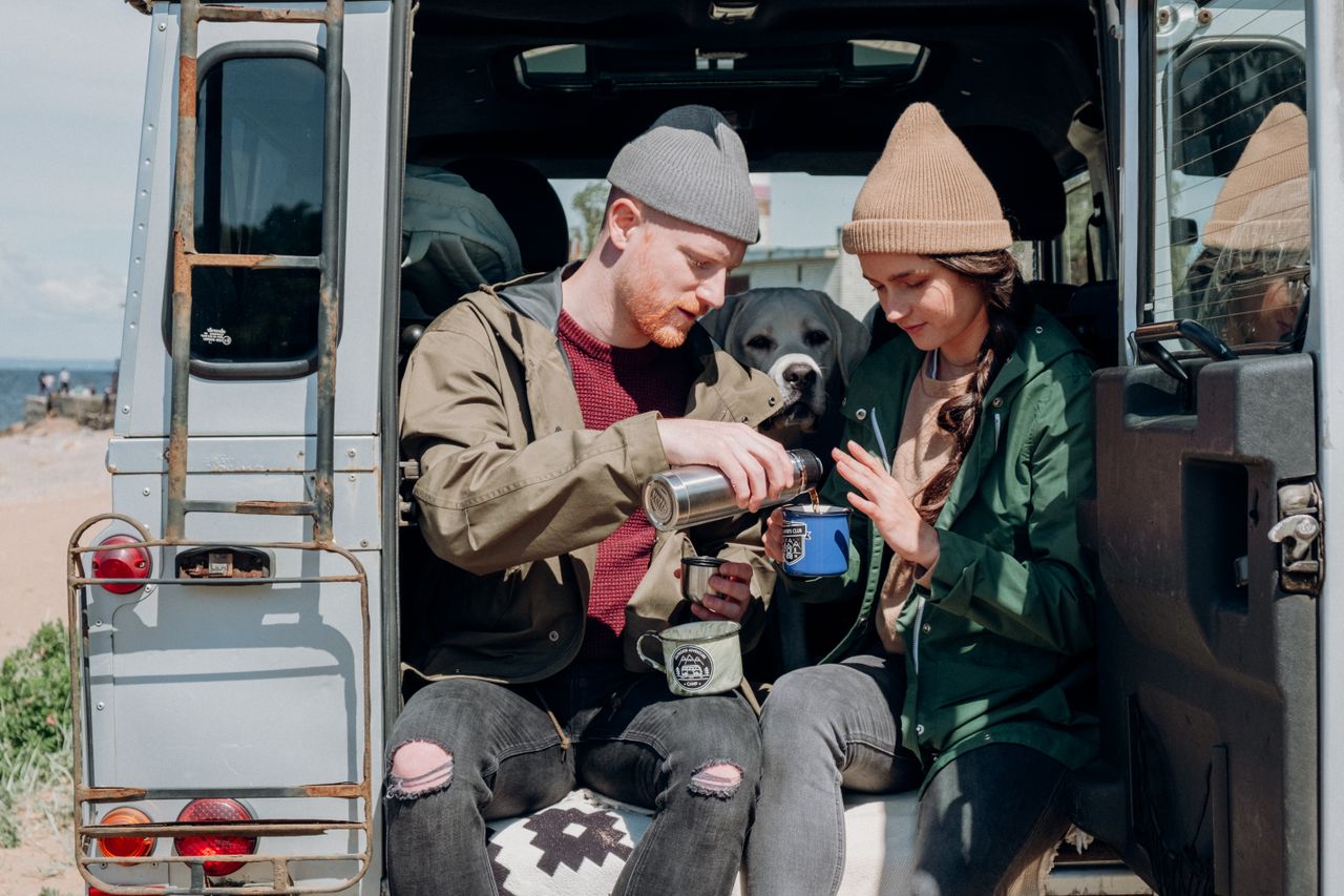 services-01 A couple pours hot drink from a thermos while sitting in a van at the beach.