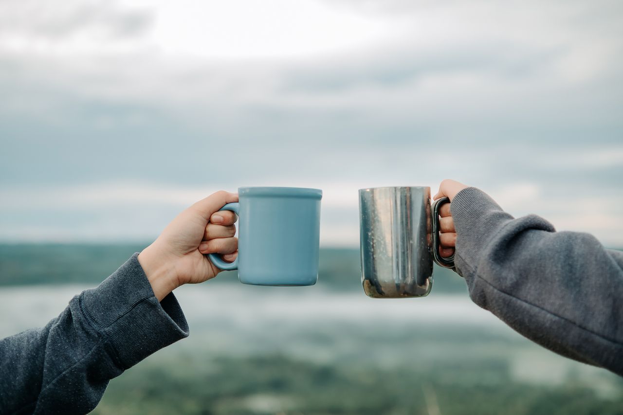 hero-img-02 Two hands holding mugs outdoors with a scenic blurry background, symbolizing friendship and relaxation.