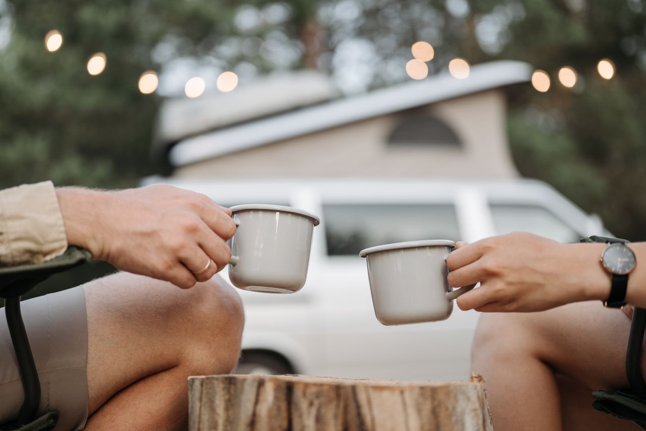 services-02 Two people enjoy a relaxing camping trip with coffee mugs, seated near a camper van under string lights.